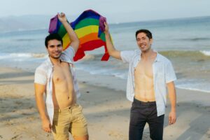 Dos hombres celebrando con una bandera arcoíris junto al mar en un día soleado