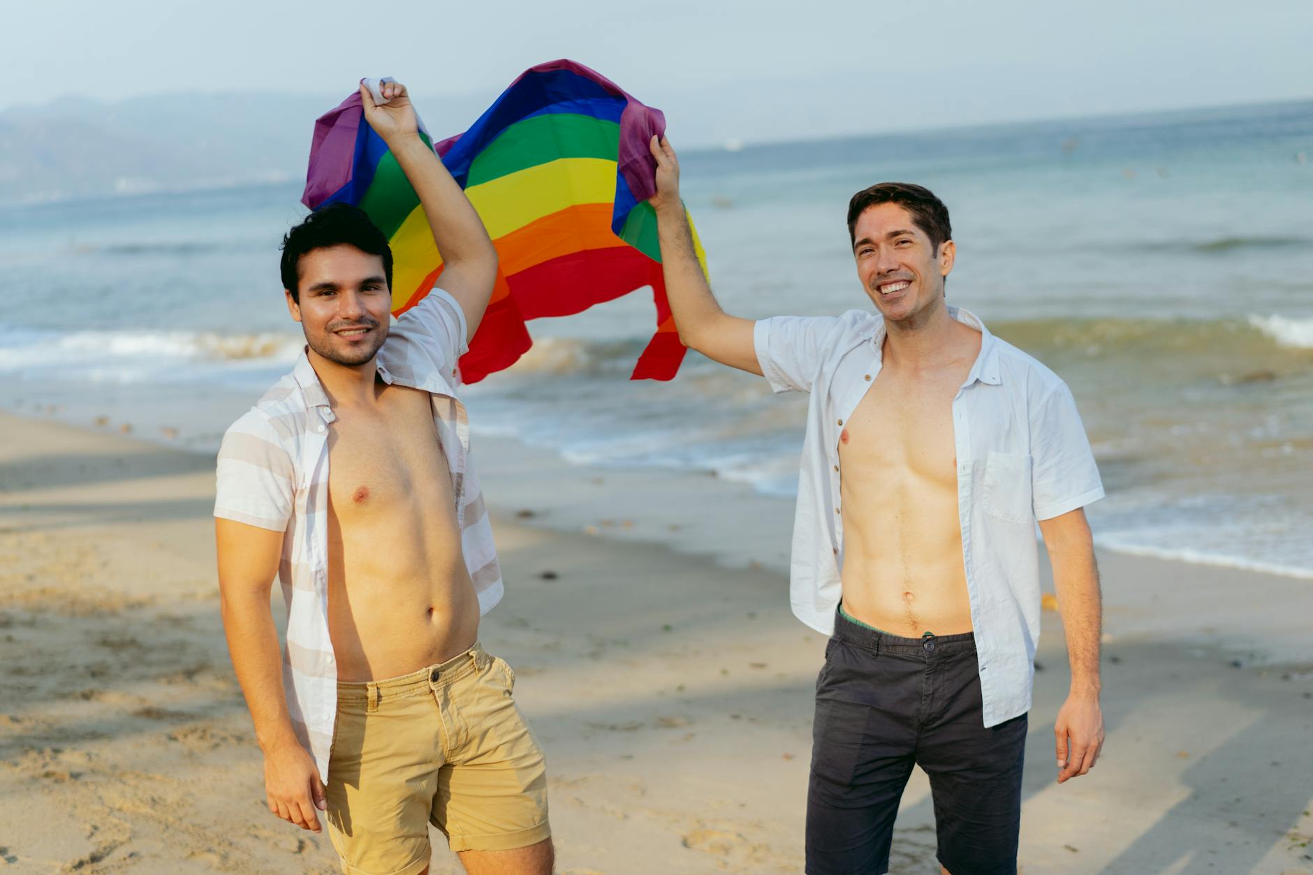 Dos hombres celebrando con una bandera arcoíris junto al mar en un día soleado