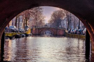 Canal de Amsterdam con barcos enmarcado por el arco de un puente, ideal para turistas jovenes