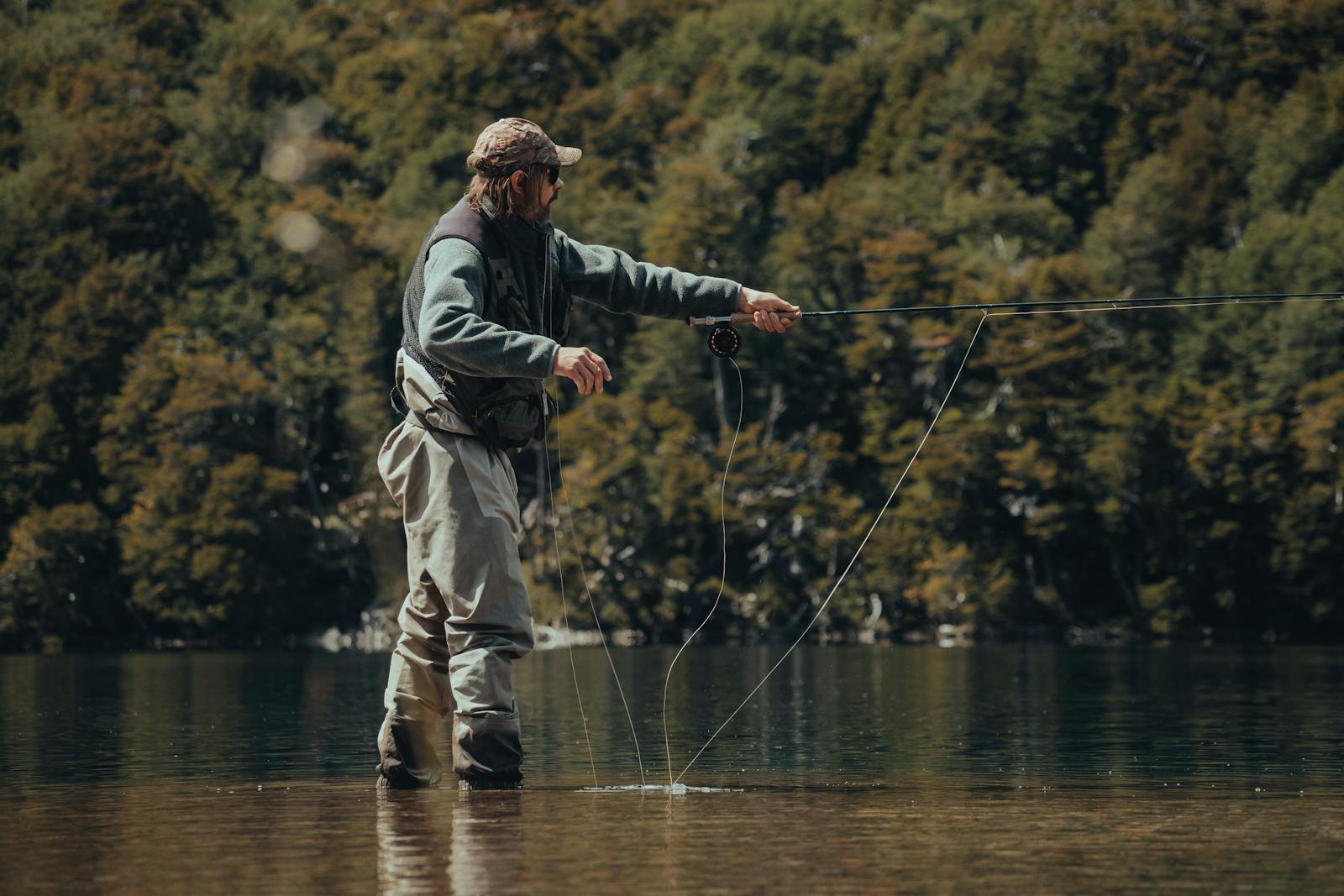 Pescador con vadeadores lanzando con cana de mosca en lago tranquilo rodeado de bosque