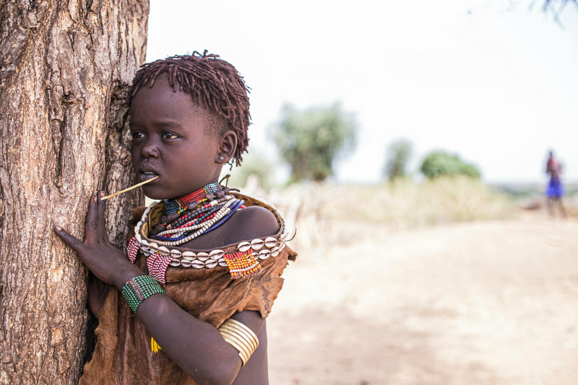 Niño etíope con vestimenta tradicional y cuentas de colores, apoyado en un árbol en el valle del Omo